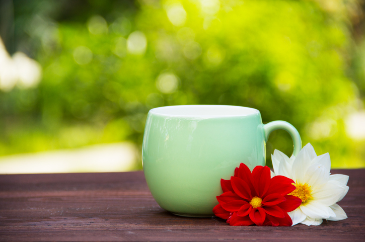 A green mug of tea sits on a wooden table outdoors. Red and white flowers lean against the cup.